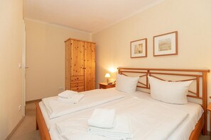 Una cama doble con ropa de cama y almohadas blancas, un armario de madera y dos cuadros en la pared.
