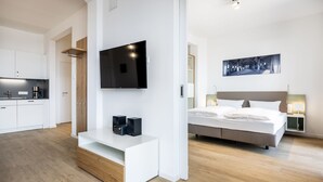 Una habitación de hotel moderna con una televisión de pantalla plana montada en la pared, una cama con cabecero, mesitas de noche con lámparas y un banco de madera con altavoces.