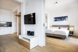 A modern hotel room with a flat-screen TV mounted on the wall, a bed with a headboard, bedside tables with lamps, and a wooden bench with speakers.