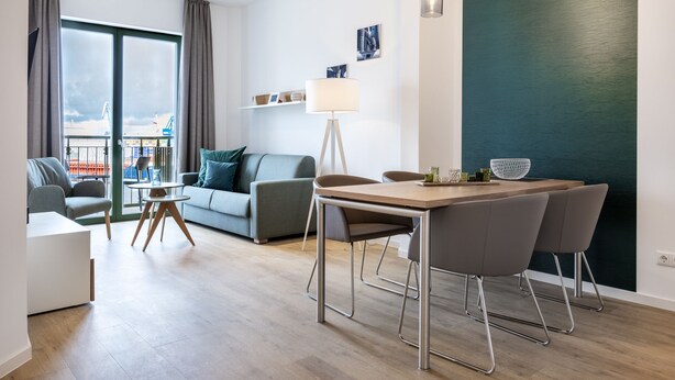 Una sala de estar moderna con sofá, una mesa de comedor con sillas, una lámpara de pie y vistas a una grúa a través de amplios ventanales.
