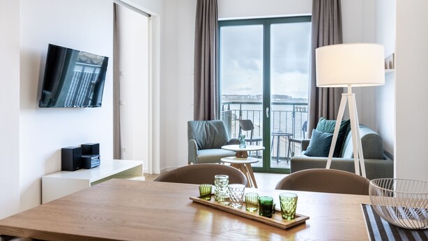 Ein modernes Hotelzimmer mit einem Holztisch, einem an der Wand befestigten Fernseher, einem Balkon mit einer Couch und Sesseln sowie einer großen Stehlampe.