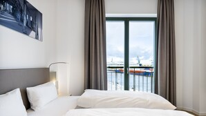 Ein Hotelzimmer mit einem großen Bett, einem Kopfteil, einem Nachttisch, einer Lampe und Blick auf einen Hafen mit Booten und Kränen.