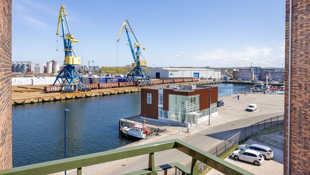 Zona industrial frente al agua, con grúas, un muelle y un edificio moderno con grandes ventanales.