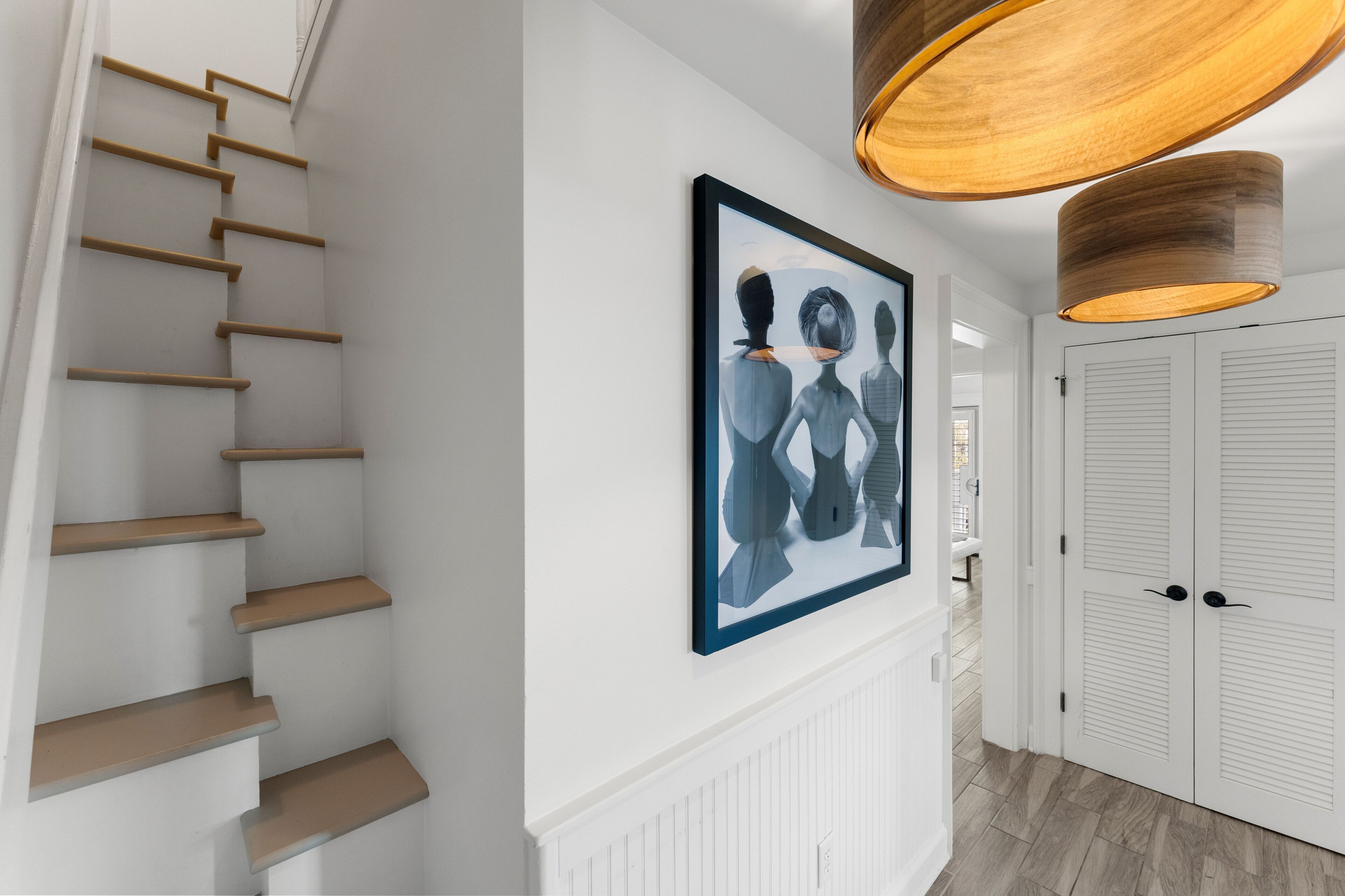 Un escalier avec des marches en bois, une œuvre d’art abstraite encadrée accrochée au mur et un luminaire suspendu en bois.