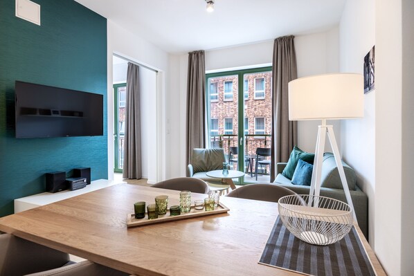 Et moderne hotelværelse med et træspisebord, et fladskærms-tv monteret på en teal farvet væg, en balkon med udsigt til en murstensbygning, og et siddeområde med en sofa og stole.