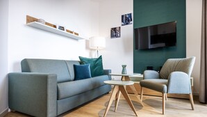 A modern living room with a grey sofa, a round coffee table, and a grey armchair. A flat-screen TV is mounted on the wall, and there is a shelf with decorative items above the sofa. - Great south-facing harbor apartment with a direct view of Wismar's old harbor (Hansestadt Wismar)