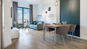 A modern living room with a sofa, a dining table with chairs, a lamp, and a view of the city through large windows.