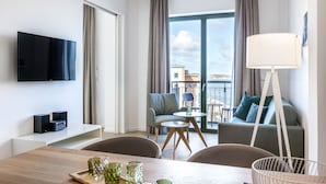 A modern hotel room with a flat-screen TV, a dining area with a wooden table and chairs, a sofa, and a balcony with a view of the city.
