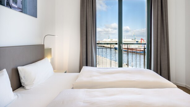 A hotel room with a large bed, a nightstand, a lamp, and a view of a port with cranes.
