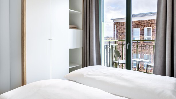 A bedroom with a bed, a wardrobe, and a balcony with a view of a brick building.