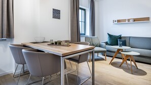 A modern living room with a wooden dining table, grey chairs, a grey sofa, and a small round table with wooden legs.