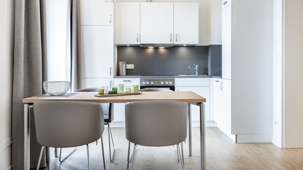 A modern kitchen with white cabinets, a wooden table, two grey chairs, and a black countertop. - Helles, Geschmackvolles Apartment in Erstklassiger Lage am Seehafen von Wismar (Hansestadt Wismar)