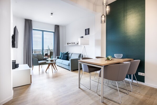 A modern living room with a sofa, a dining table with chairs, a TV, and a large window with curtains.