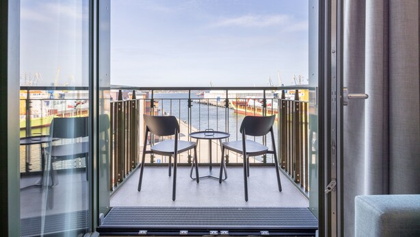 Un balcón con dos sillas y una mesa pequeña, con vista a un puerto con grúas y edificios.