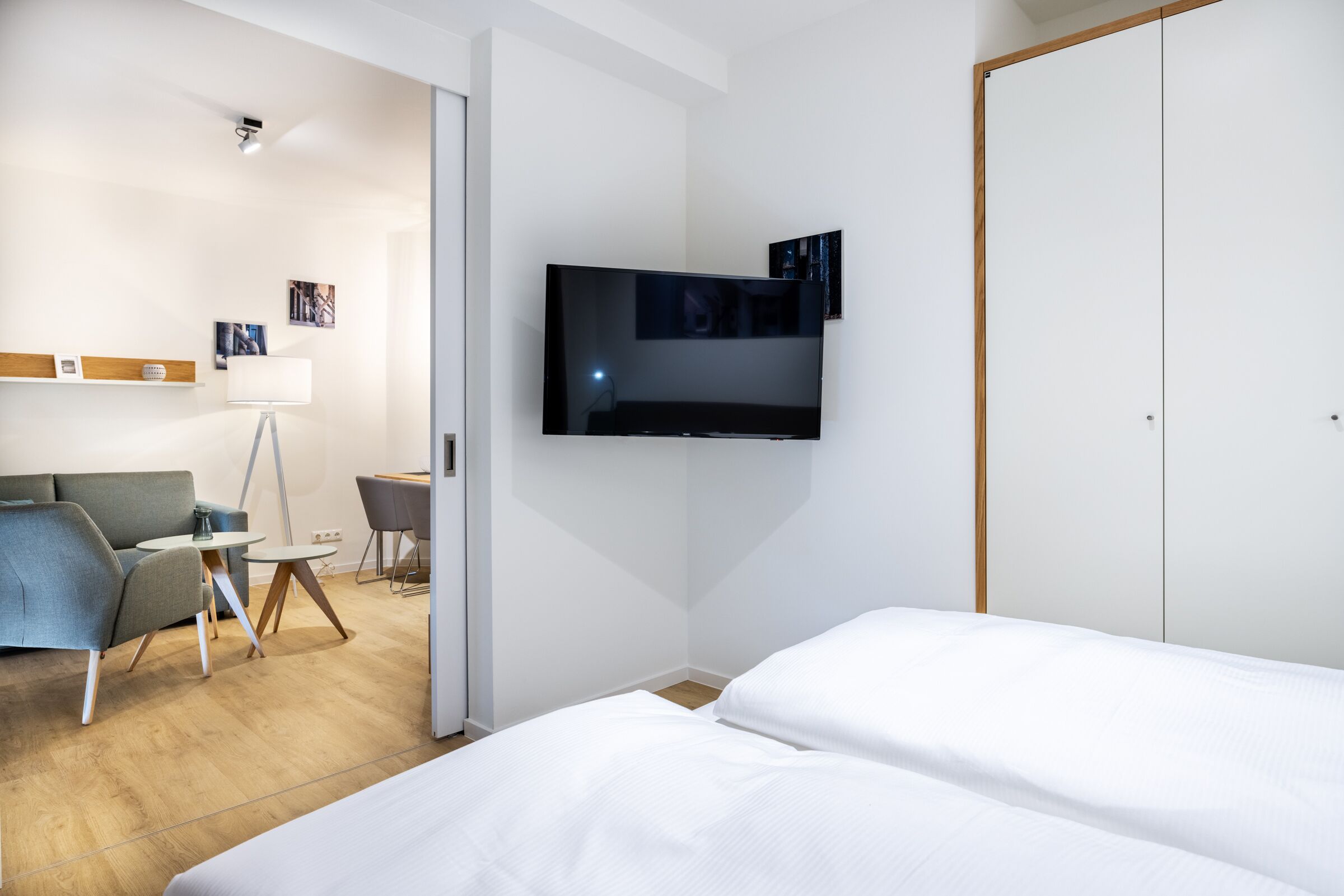 A modern hotel room with a bed, a TV mounted on the wall, a sofa, a small table, and a chair.