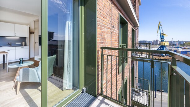 A modern balcony with a view of a river and construction cranes, a glass door leading to a kitchen area with white cabinets and a dining table.