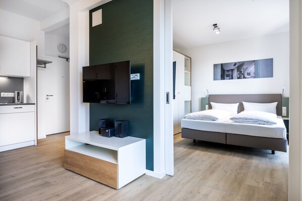 Quarto de hotel moderno com uma cama, uma TV fixada na parede, uma pequena escrivaninha e uma área de cozinha visível por uma porta aberta.