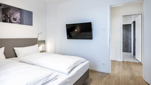 A modern hotel room with a large bed, a flat-screen TV mounted on the wall, and a wooden floor.
