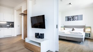 A modern hotel room with a flat-screen TV, a bed, bedside lamps, a small desk with speakers, and a kitchen area in the background.