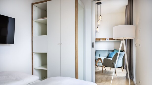 A modern hotel room with a large bed, a wardrobe with multiple shelves, a desk with a chair, and a large floor lamp.