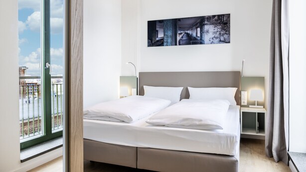 A modern hotel room with a large bed, a view of the city through a large window, and a painting on the wall.