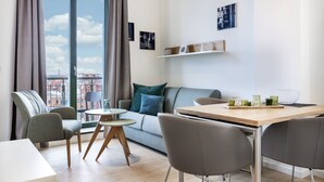 A modern living room with a sofa, a dining table, and chairs. There is a window with curtains, a small round table, and a shelf with decorative items.