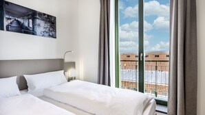 A hotel room with two beds, a large window with a view of buildings, and a painting on the wall.