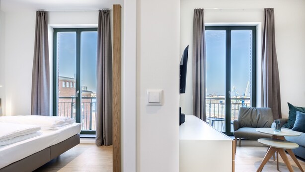 Una habitación de hotel moderna con cama, zona de comedor con sofá y mesa, y ventanas amplias con cortinas.