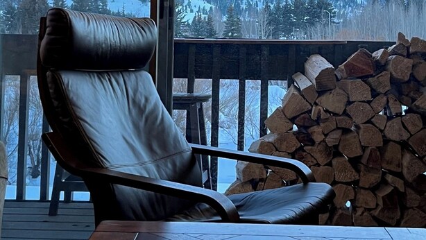A room with a large window, a leather chair, a table with a book, and a stack of firewood. - Snowcreek Resort Vacation Rental 99 (Mammoth Lakes)