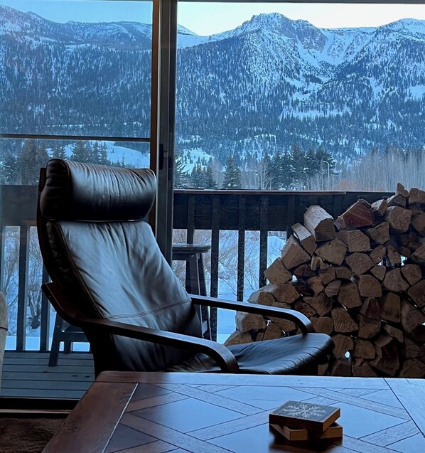 A room with a large window, a leather chair, a table with a book, and a stack of firewood. - Snowcreek Resort Vacation Rental 99 (Mammoth Lakes)