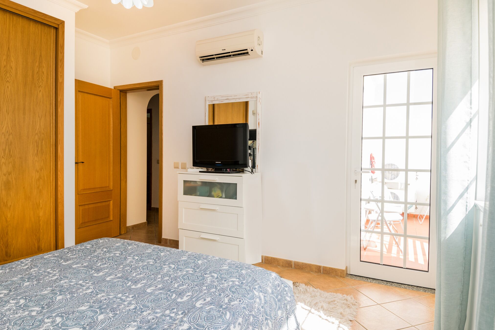 Un dormitorio con cama, televisor sobre un mueble blanco, puerta y ventana con vista a muebles de exterior.