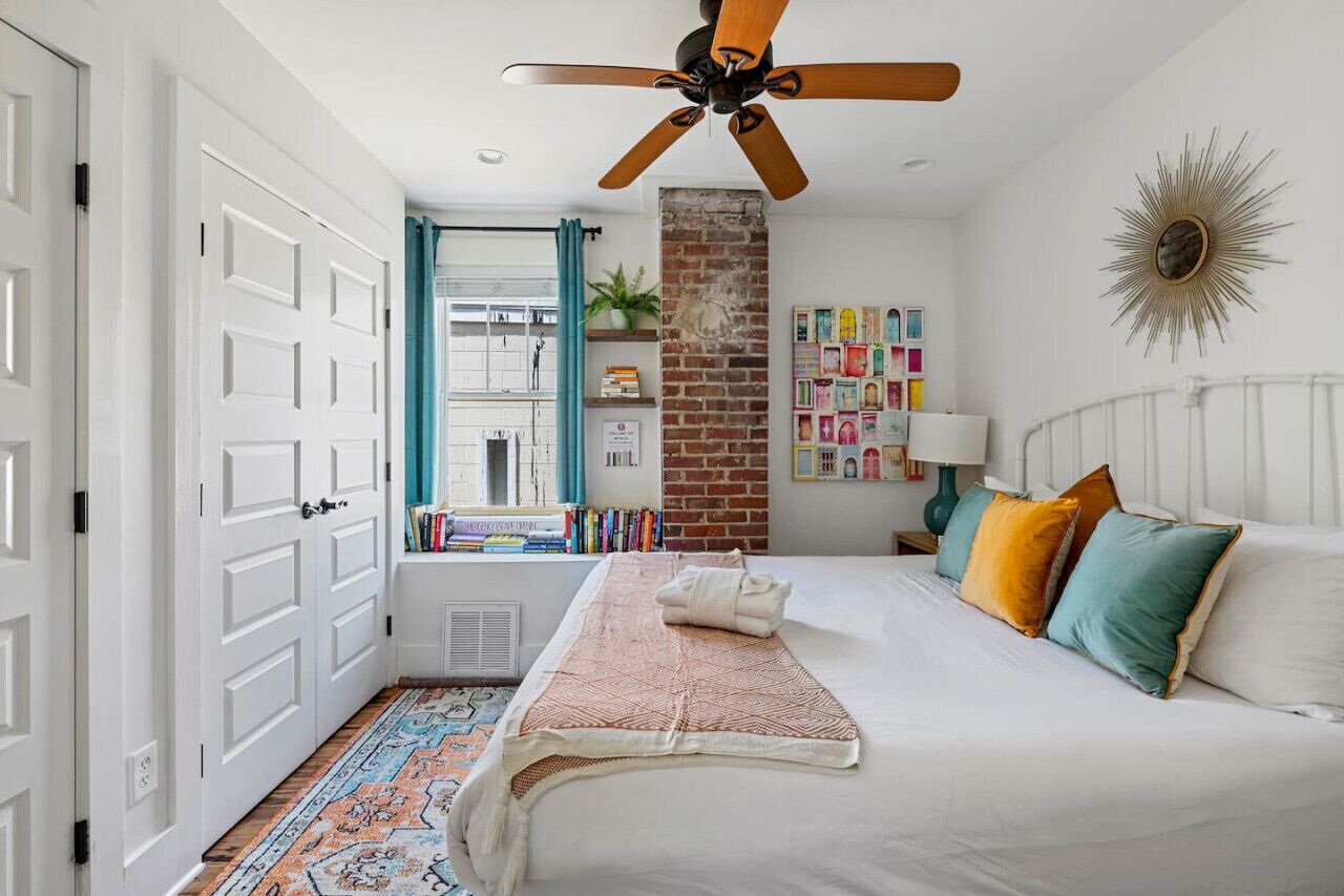 Une chambre avec un lit, un ventilateur de plafond, un mur en briques, une fenêtre avec des rideaux, une bibliothèque et une décoration murale en forme de soleil rayonnant.