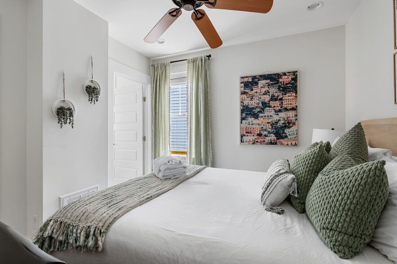 Une chambre à coucher avec un lit, un ventilateur de plafond, une fenêtre avec des rideaux, une plante fixée au mur et un tableau représentant un paysage urbain.