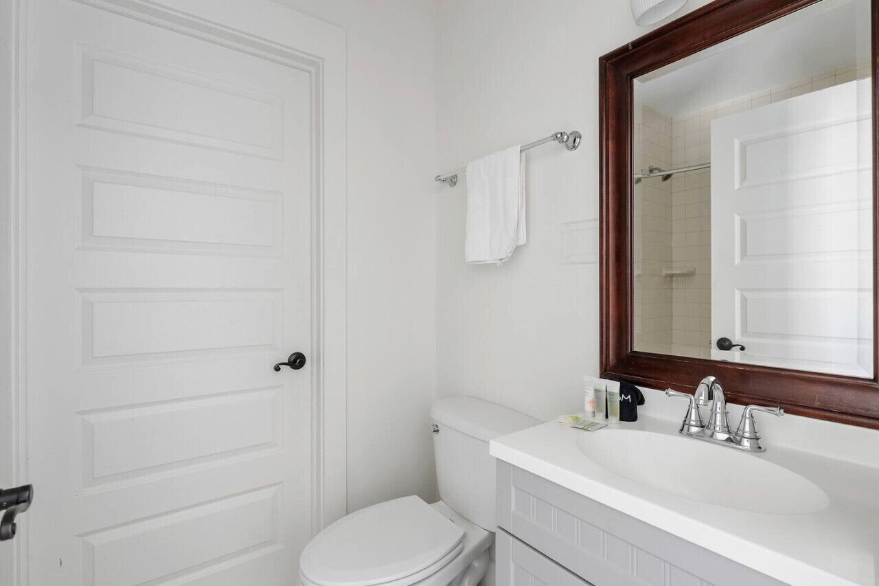 Une salle de bain avec des toilettes blanches, un lavabo blanc surmonté d’un grand miroir et un porte-serviettes.
