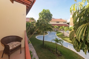 A balcony with a wicker chair, a swimming pool, and surrounding greenery.