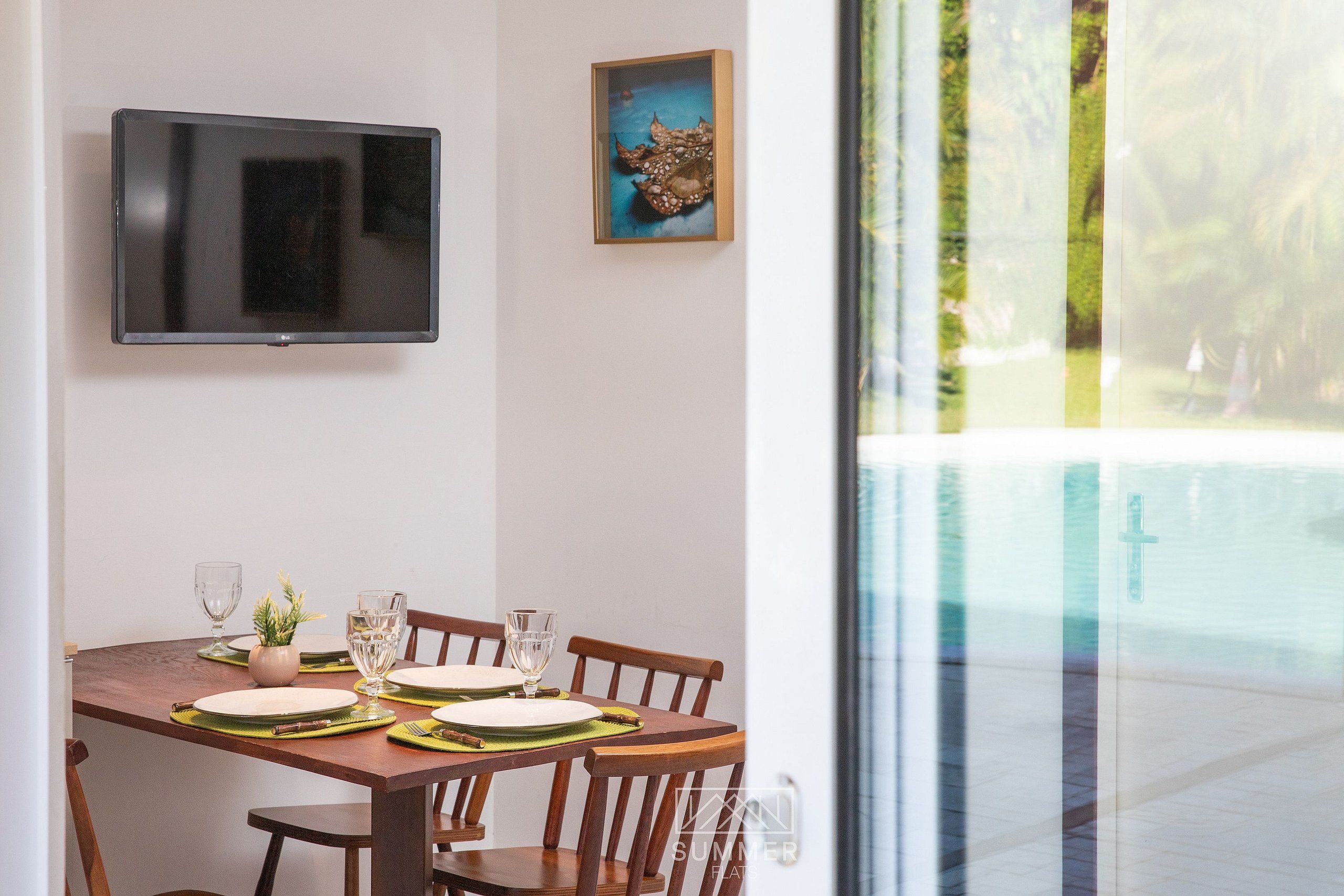 A dining area with a table set for four, a television mounted on the wall, and a window with a view of greenery.