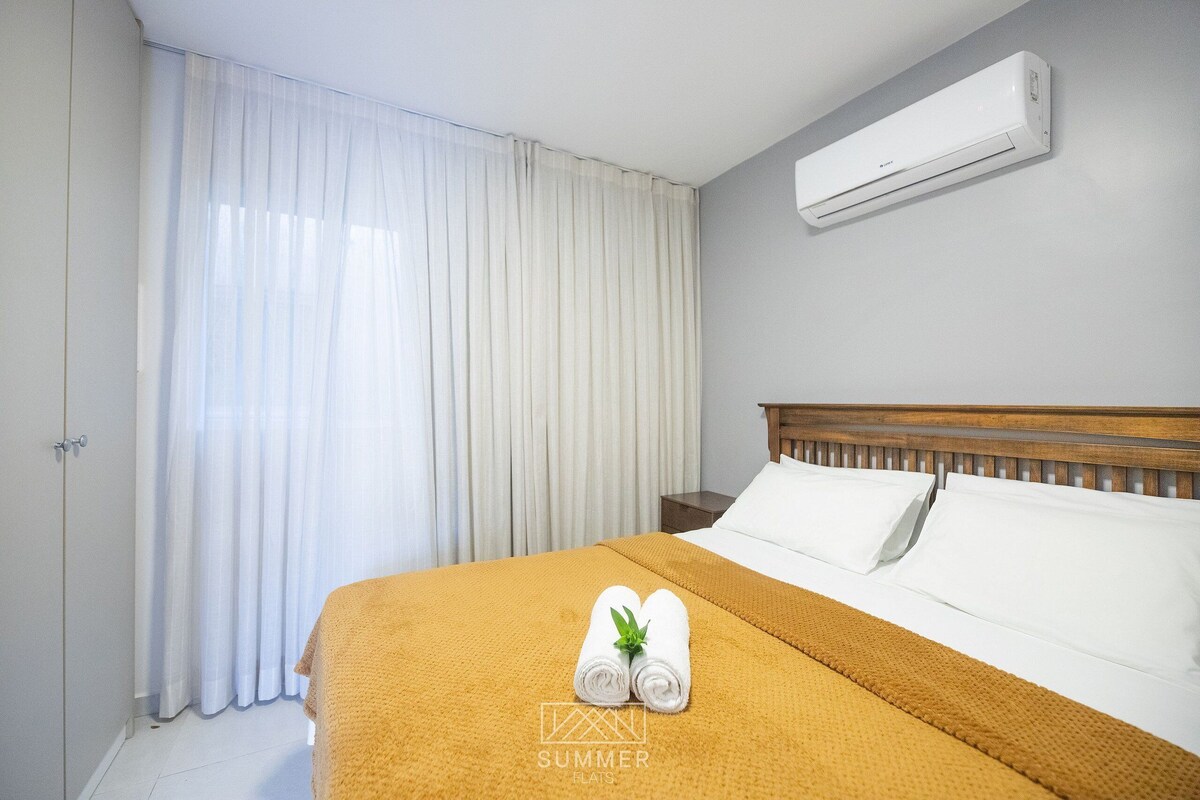 A hotel room with a bed, a nightstand, a window with curtains, and an air conditioning unit.
