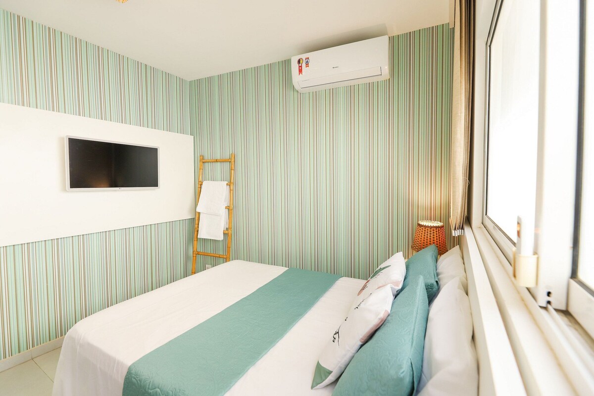 A hotel room with a bed, a flat-screen TV, an air conditioning unit, a window with a view, and a striped wall.