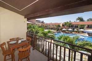 Un balcón con mesa y sillas de madera, con vista a una piscina con sombrillas y palmeras.