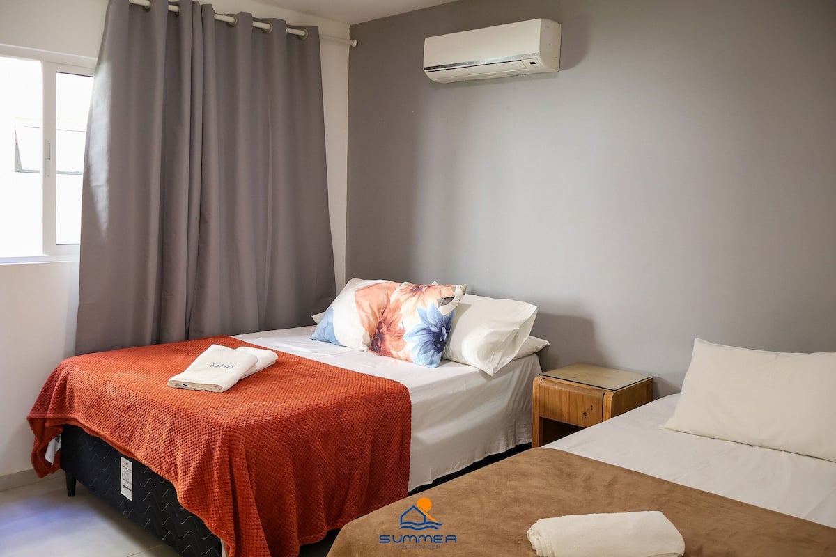 Una habitación de hotel con cama, mesita de noche, aire acondicionado y ventana con cortinas.