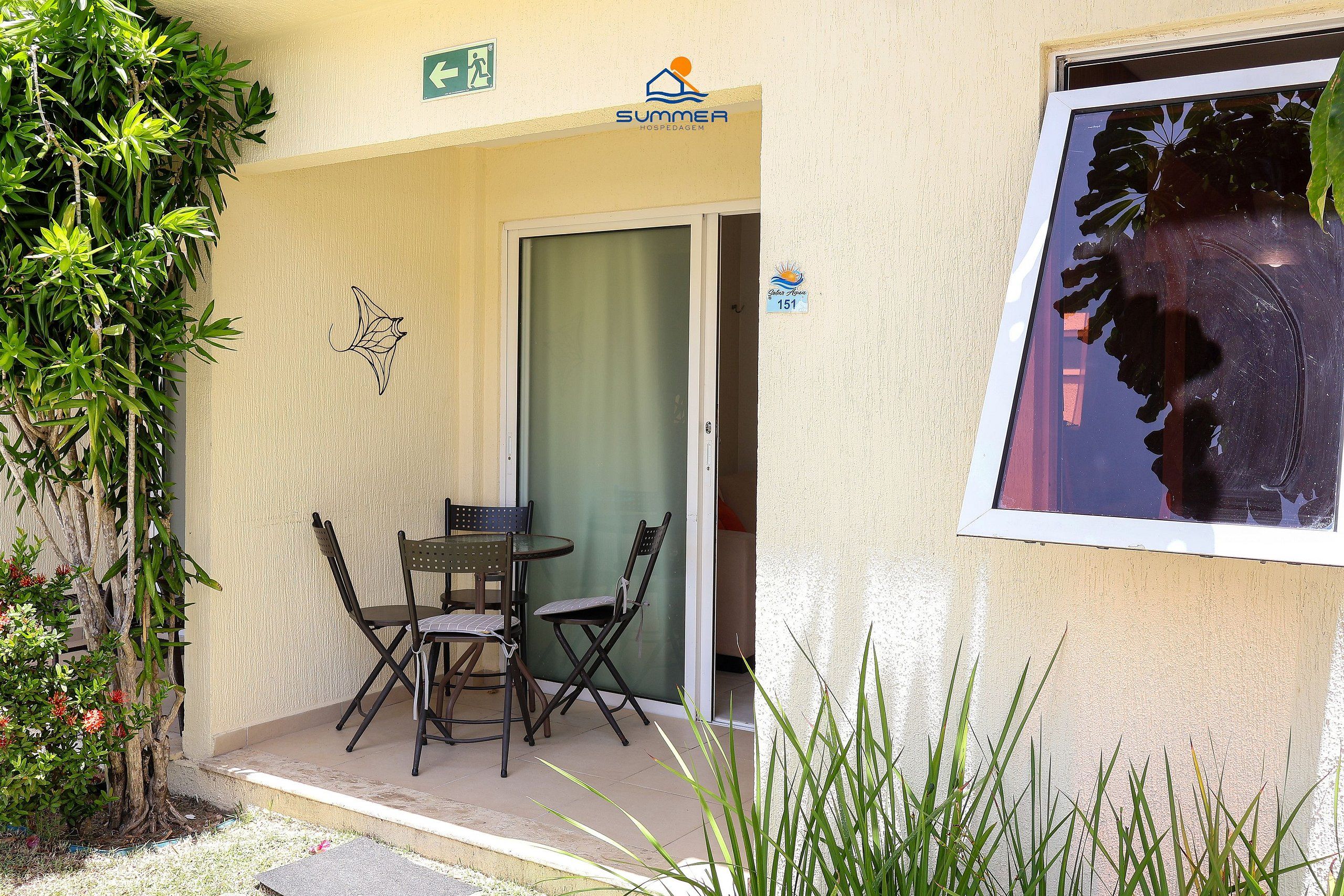 En byggnadsentré med en glasdörr, ett litet utomhusområde med bord och stolar, och en skylt som visar namnet ”Summer Resort”.