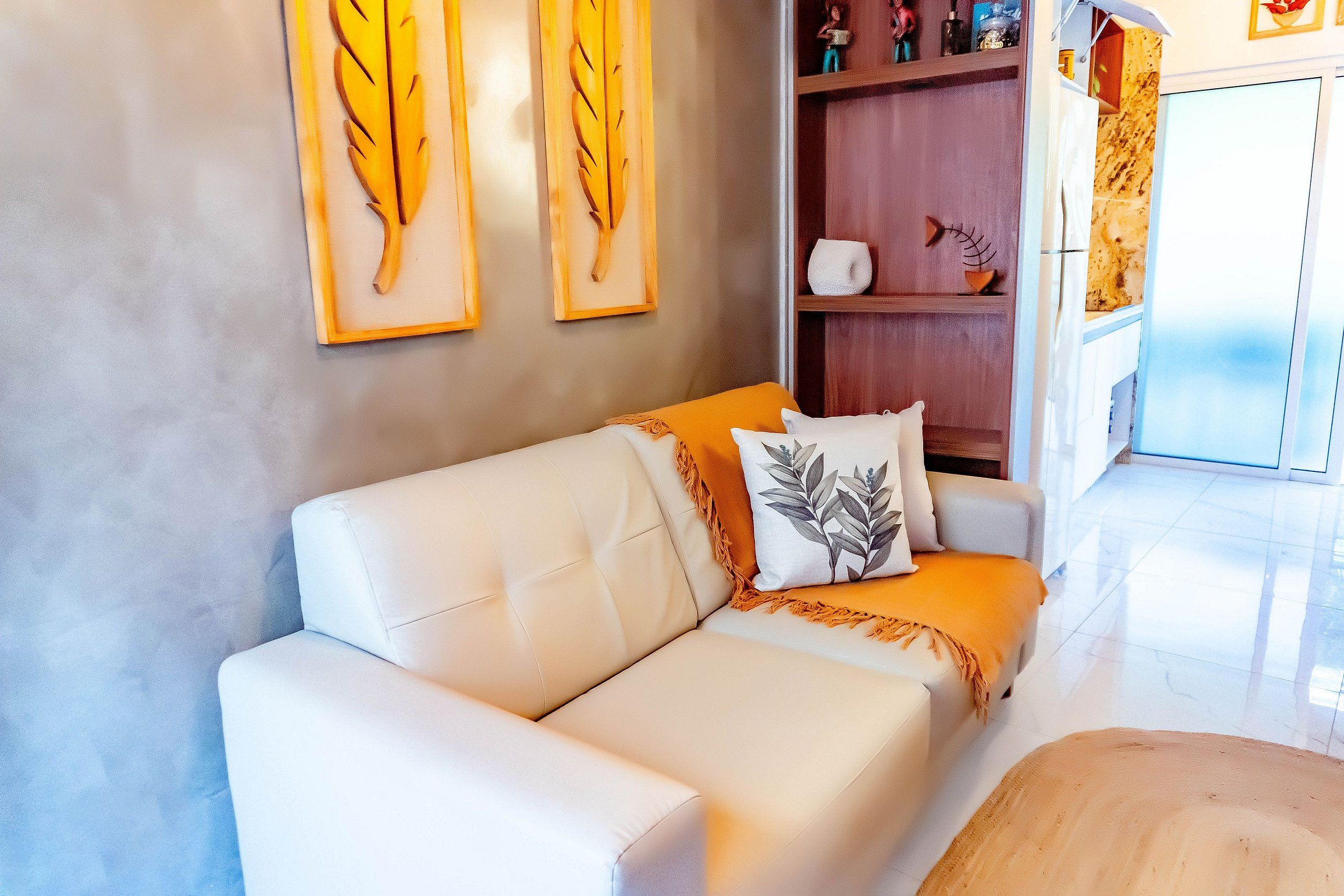 A modern living room with a white leather sofa, a decorative pillow, and two wall-mounted leaf paintings.
