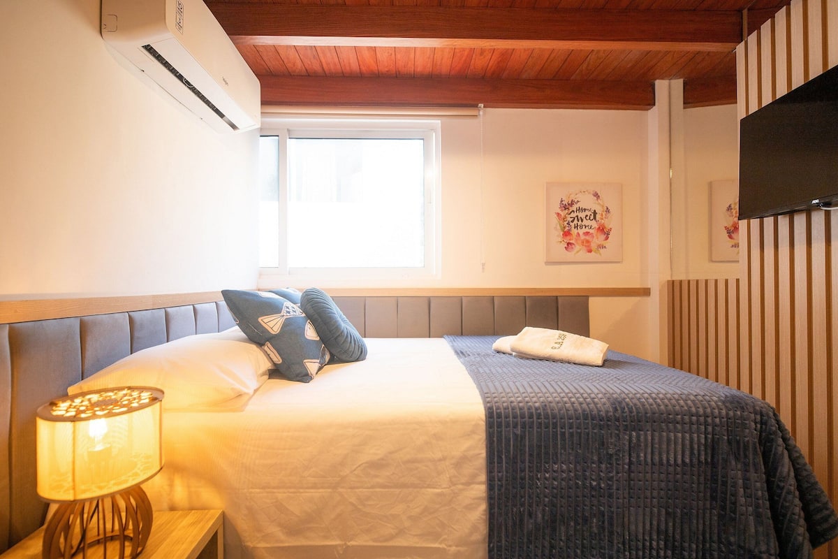Habitación de hotel con cama, mesita de noche con lámpara, ventana y televisor montado en la pared.