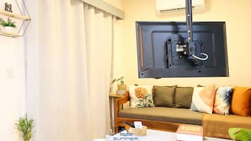 A living room with a sofa, a coffee table with a book and a basket, a mounted TV, and a wall-mounted air conditioner.