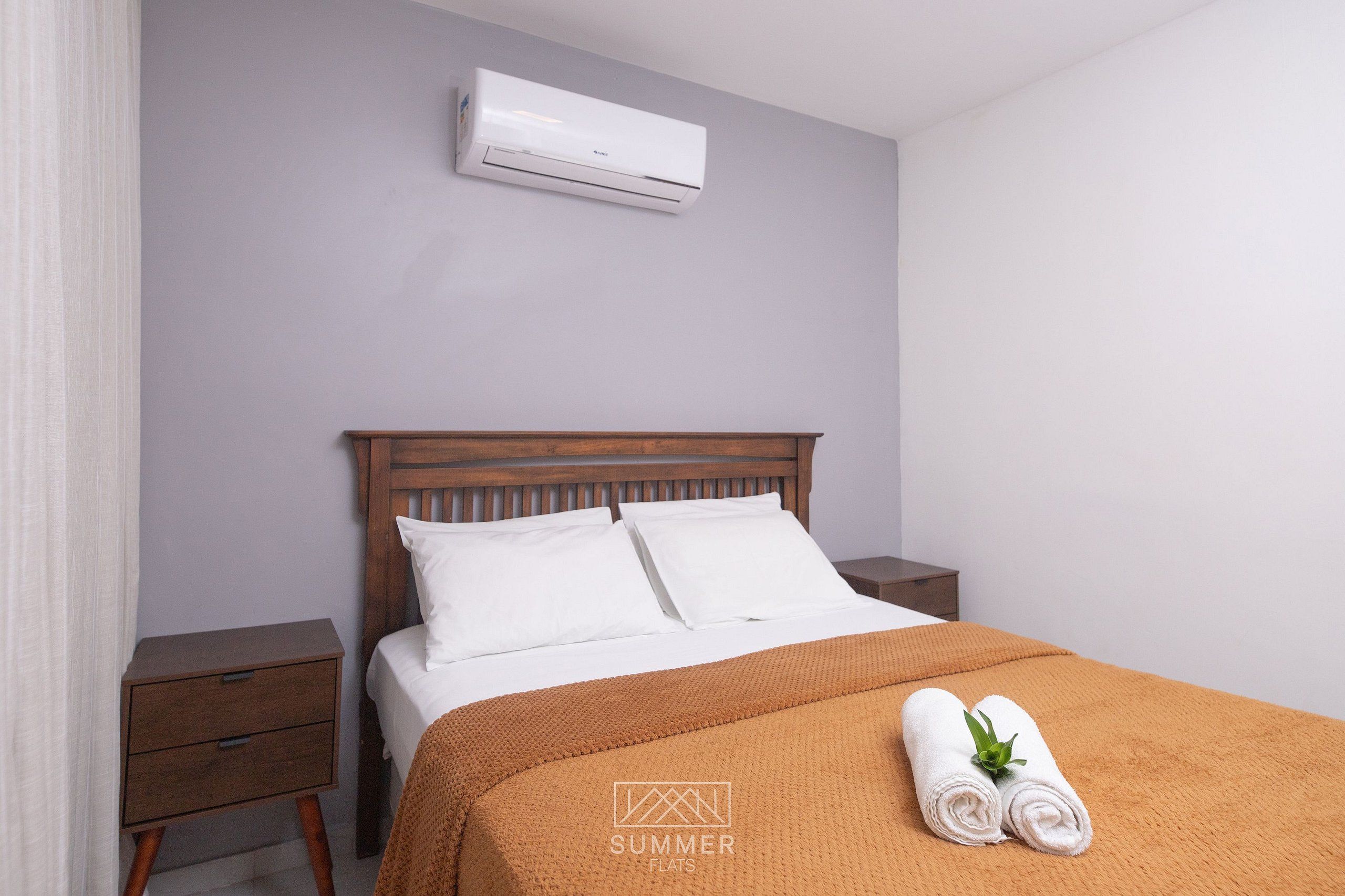 A bedroom with a bed, a nightstand, a wooden headboard, a white air conditioner, and a rolled towel on the bed.