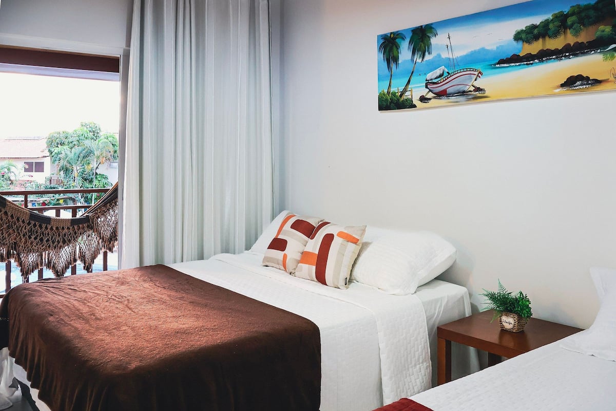 Un cuarto de hotel con una cama, una mesita con una planta, un cuadro de una playa y vistas al exterior a través de una ventana.