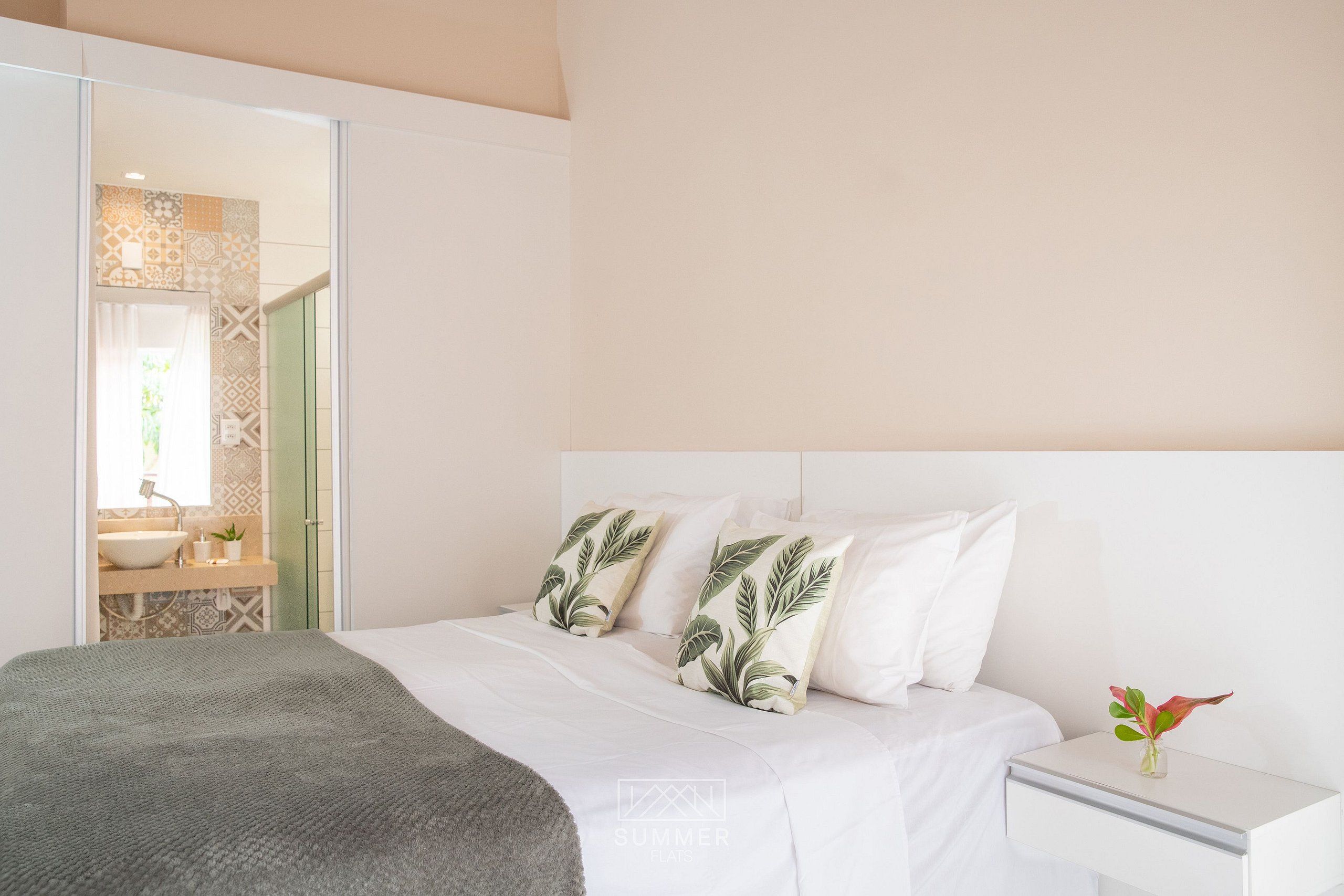 Un dormitorio con cama, almohadas, una mesita de noche y una planta pequeña.