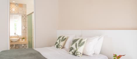 Un dormitorio con cama, almohadas, una mesita de noche y una planta pequeña.