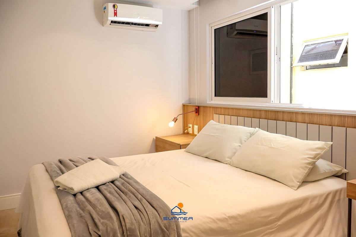 Un dormitorio con cama, mesita de noche, aire acondicionado, ventana con vistas y una lámpara de pared.