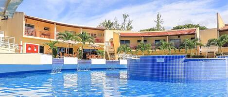 Uma piscina com água azul cristalina, área de deck com azulejos e um edifício com várias varandas, com palmeiras ao fundo.